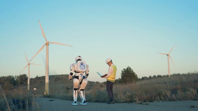 A human-like robot and a male operator are walking along the set of windmills