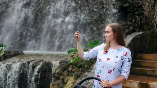 A young mother walking with her baby near the waterfall takes pictures on her smartphone while traveling with her family.