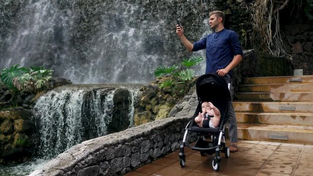 A young father walking with a child in a wheelchair near the waterfall takes pictures on his smartphone for social networks and posting on the Internet while traveling with his family.