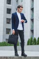 Business style. Young businessman in his hand the briefcase. The guy in the blue jacket looks at his watch.