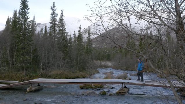 A man traveler crosses a mountain stream over a bridge. Beautiful view. He photographs the landscape on a smartphone.
