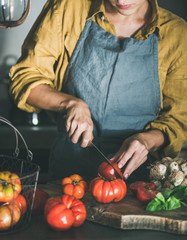 Woman in grey linen apron cutting and cooking tomato sauce, canned tomatoes or pasta with heirloom tomatoes, basil and garlic at kitchen counter. Seasonal healthy cooking, slow food or comfort food