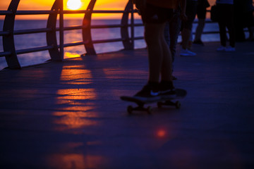 People walk along the promenade in the yellow and orange rays of the sun at sunset. Go on foot, ride a skateboard, silhouettes in the light of the sun and the shadows from the fence and railing. 