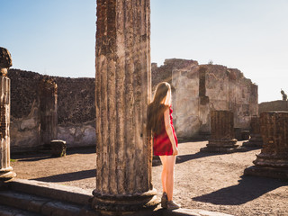 Fototapeta premium Stylish woman in a red dress looking into the distance against the backdrop of ancient ruins and the sun's rays of the evening sun. Pompeii, Italy. Concept of travel and leisure.
