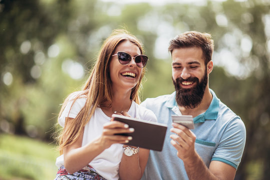 Cheerful young couple sitting on a park bench and uses a digital tablet for online shopping