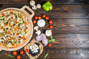 Food background design. Big pizza with cheese, tomatoes, black olives and paprika on a round cutting board on a dark wood. Ingredients.