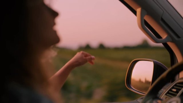 Cinematic inspirational video of young woman travelling by car or camper van, opens window to breathe fresh air of countryside, moves hand in wind. Sings melody of song, summertime vacation vibes