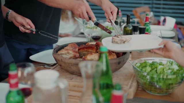 Man cuts down and serves delicious, juicy meat pieces from barbecue into empty white plate for guests or friends. Beautiful table with fresh and tasty food, salads and drinks. Summer holiday concept