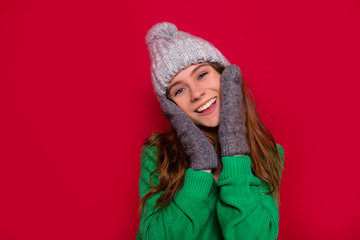 Studio shot of happy smiling girl with adorable smile and blue eyes touching her face dressed winter cap and mittens on isolated red background