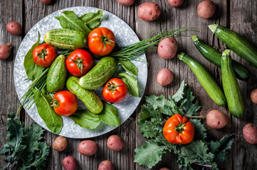 A produce scene of freshly harvested cucumber, tomatoes, red potatoes, kale, zucchini squash and kale on a rustic background. 