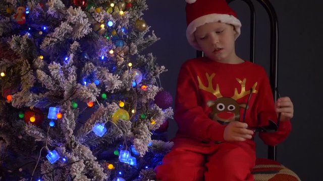 Cute boy from school for visually impaired tries on glasses with patch for treatment of strabismus. child is dressed as Santa Claus deer and is rehearsing Christmas show next to Christmas tree