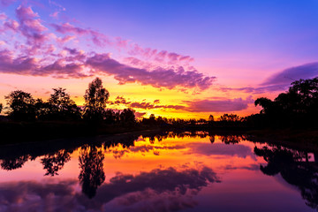Sun setting with silhouette trees and bush around the big pond in rural of Thailand, shot taken by using long exposure therefore smooth water surface like mirror