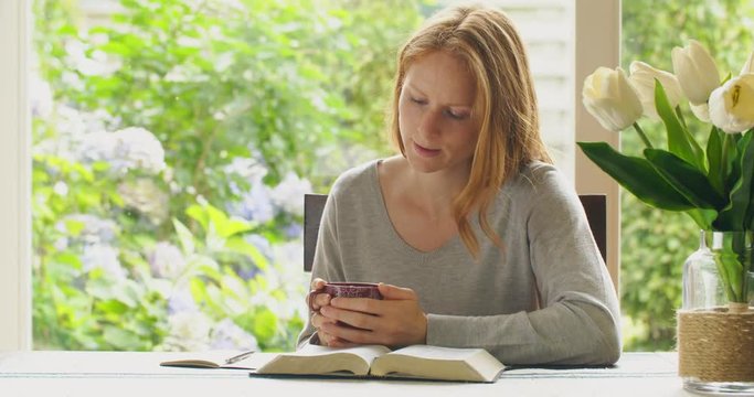 Young Christian woman reading the Bible and drinking coffee at home.