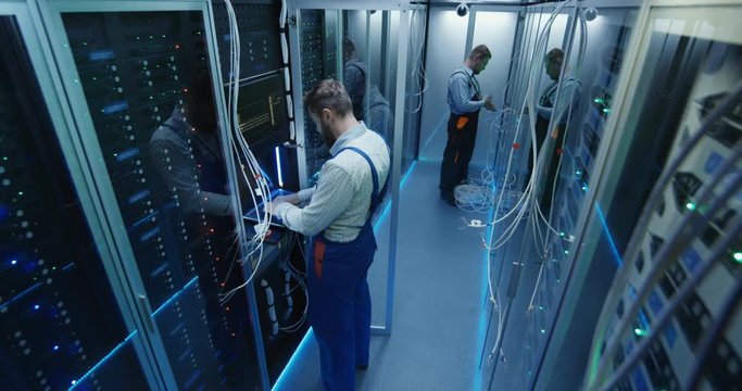 Wide shot from above like from surveillance camera of people working in a data center server room with rows of server racks checking the equipment and discussing their work.