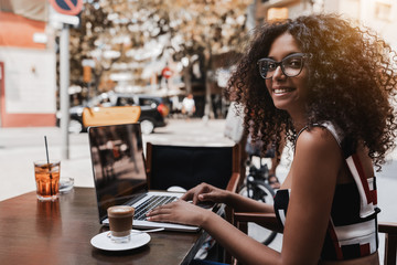 Smiling young ravishing Brazilian businesswoman in eyeglasses and with a gorgeous curly bulky hair is sitting in a street bar, working on her laptop and drinking delicious cappuccino from the cup near - obrazy, fototapety, plakaty