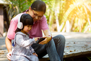 Asian smart mother teaches or suggests her little daughter to use smartphone and headphones for listen nursery songs or watch kids movie on the wooden bridge at the public garden or the nature forest.
