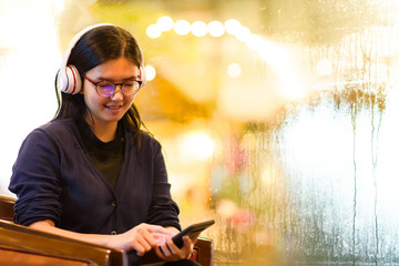 Asian charming beautiful happy business woman or student listening or watching a content or a movie or a video from smartphone and headphones in coffee shop or smart office on raining day night.
