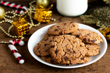 Chocolate chip cookies freshly baked on white plate with glass bottle of milk and gift festive decoration on wooden table background. Christmas cookies. Homemade pastry.