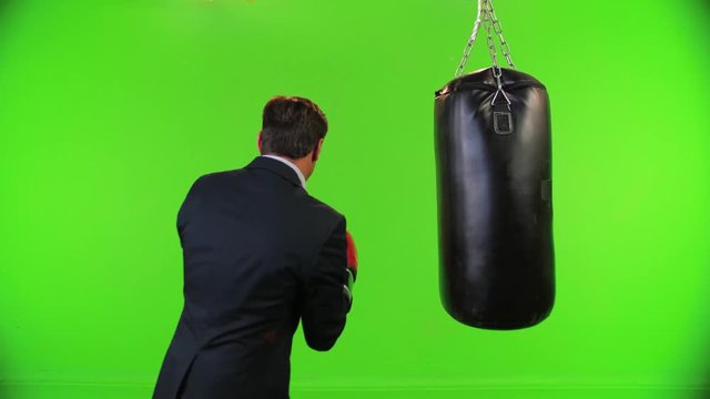 Businessman with boxing gloves trains from the back, swings and hits, looking forward over a green screen,  medium shot.
