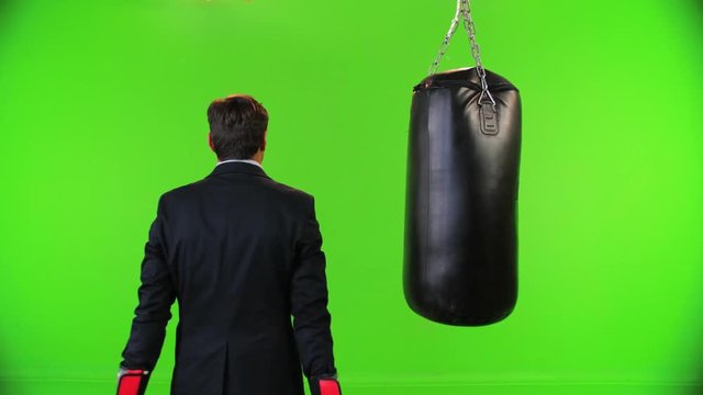 Businessman with boxing gloves trains from the back, swings and hits, looking forward over a green screen,  medium shot.