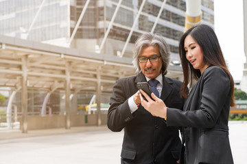 Asian smart old manager boss point on his secretary smartphone for tell her about the company project and his schedules and appointments at skywalk on metro station in center business zone of city.