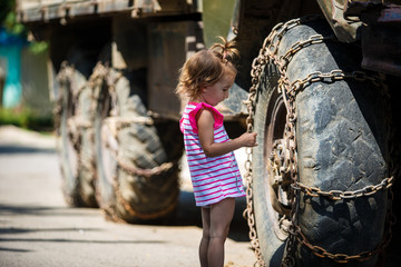 Fototapeta na wymiar A little girl in a pink dress next to the big wheels of a car in chains. The contrast is a nice girl and huge wheels of the old ZIL
