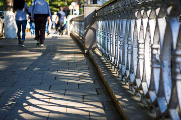 Walking along the sidewalk in Borjomi, Georgia. The pattern of chiaroscuro from the railing on the tile sidewalk, people walking on the road in bright sunlight