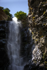 Obraz premium Gveleti waterfall in Georgia. The stream of water pouring from the top on the wet gray rocks, the sunlight playing in the spray of water and is reflected on the wet black rocks.