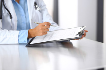 Unknown doctor woman filling up medical form while sitting at the desk in hospital office, close-up...