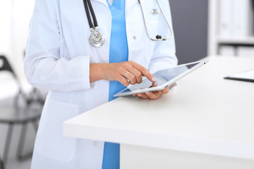 Close-up of female physician using digital tablet  while standing near reception desk at clinic or emergency hospital. Unknown doctor woman at work. Medicine and healthcare concept