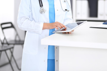 Close-up of female physician using digital tablet  while standing near reception desk at clinic or emergency hospital. Unknown doctor woman at work. Medicine and healthcare concept