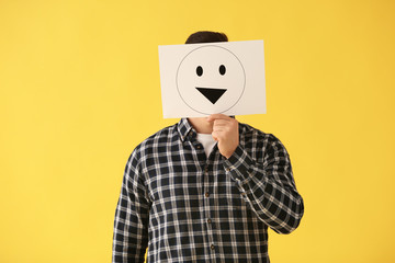 Man hiding face behind sheet of paper with drawn emoticon on color background