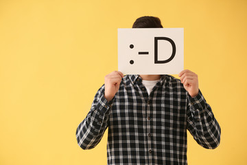 Man hiding face behind sheet of paper with drawn emoticon on color background