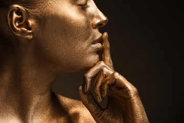 Beautiful young woman with golden paint on her body showing silence gesture against dark background