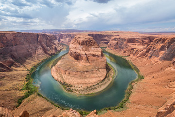 Horseshoe Bend view from above to Glen Canyon, green meander of Colorado river bends around red rock formation, storm clouds in the background. Wide angle landscape.