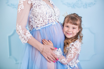 Fototapeta na wymiar Beautiful pregnant woman wearing long blue luxury dress with daughter posing in studio with happy lovely faces with brunette curly hairs