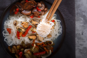 Traditional wooden chopsticks with coiled rice noodles on the background of a dish with seafood, vegetables and peppers. Close-up. Top view