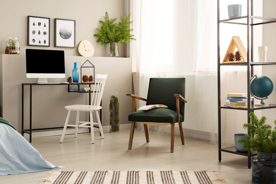 Open book on dark green retro armchair in the middle of stylish workspace with metal shelf with books and desk with all in one computer