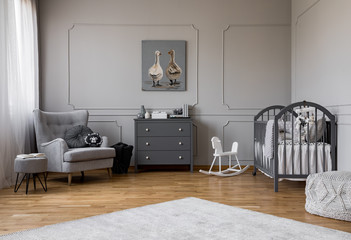 Grey chest of drawers in the middle of elegant grey baby room with comfortable armchair and wooden...