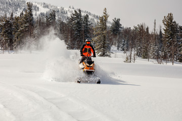 the guy rides a snowmobile in a mountain valley on the background of the winter forest, leaving behind a trail of splashes of white snow. bright snowmobile and suit without brands. very high quality