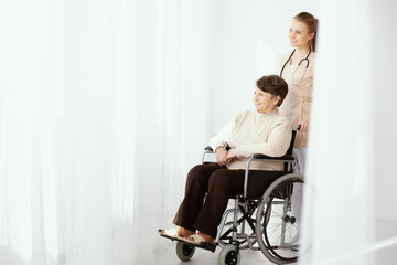 Smiling caregiver supporting paralysed elderly woman in the wheelchair next to copy space