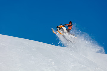 Naklejka na ściany i meble the guy is flying and jumping on a snowmobile on a background of blue sky leaving a trail of splashes of white snow. bright snowmobile and suit without brands. extra high quality 
