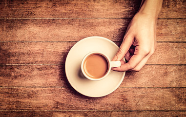 Female hand holding white cup of coffee wiht milk on wooden table. Above view