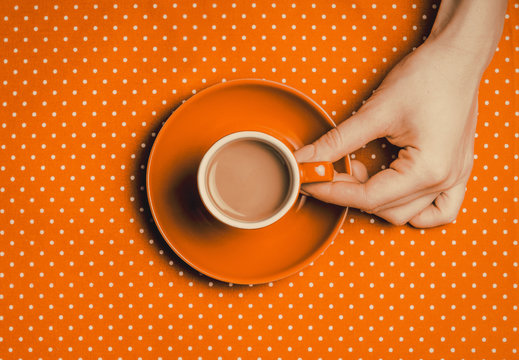 female hand holding an orange cup of coffee with milk on polka dot background. Above view