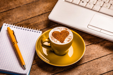 green cup of coffee with cream heart shape near laptop and pen with notebook on wooden table.