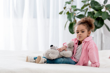 Upset  african american kid sitting on the bed with teddy bear on her knees
