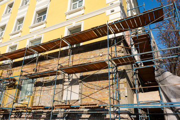 Iron and wooden scaffolding near the yellow house during reconstruction, building renovation during daytime. building exterior, construction and repair industry, yellow wall and windows.