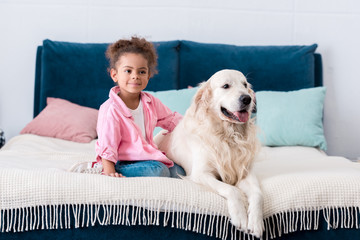 Cute african american child sitting on the bed with her dog beside