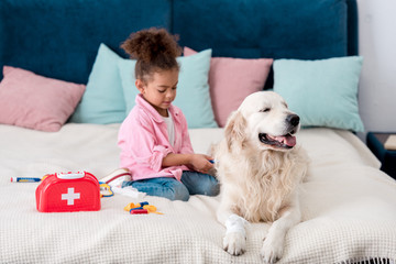 Cute african american child sitting on the bed with toys near her dog