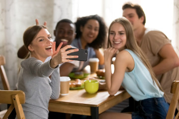Cheerful girl sitting with diverse mixed race friends around table in cafe holds mobile phone take selfie photo for sharing on social networking services close up focus on female hand and smartphone.